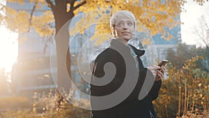 Portrait of young caucasian woman using phone in the city park in front of office building in autumn