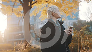 Portrait of young caucasian woman using phone in the city park in front of office building in autumn