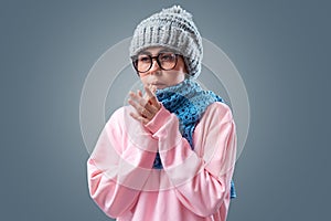 Portrait of young caucasian woman wearing glasses, in hat and knitted blue scarf is warming herself. Gray background