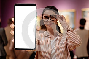 Portrait of young Caucasian woman wearing glasses shows cellphone with mock up. Concept of cultural education and Museum photo