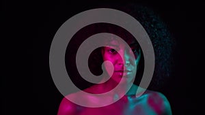 Portrait of young charming african girl in pink neon light at black background. Tempting woman with perfect makeup