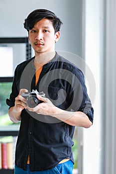 Portrait of a young cheerful asian professional male photographer standing near window in a modern office, posturing by holding a