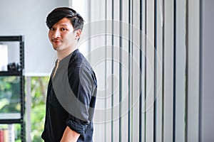 Portrait of a young cheerful asian professional male photographer standing near window in a modern office, posturing by holding a