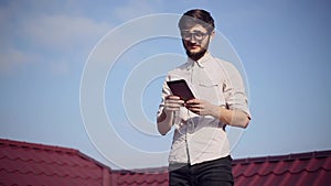 Portrait of young cheerful hipster on house roof with smartphone and earphones listen the music, wearing round sunglasses.