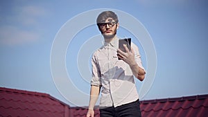 Portrait of young cheerful hipster on house roof with smartphone and earphones listen the music, wearing round sunglasses.