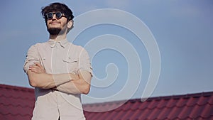 Portrait of young cheerful hipster on house roof with smartphone and earphones listen the music, wearing round sunglasses.