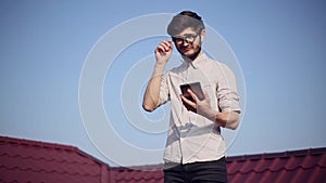 Portrait of young cheerful hipster on house roof with smartphone and earphones listen the music, wearing round sunglasses.