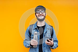 Portrait of young cheerful hipster man on orange background. Holding steel thermo eco bottle for water and smartphone.