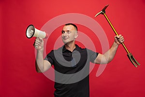 Portrait of young cheerful man with pickaxe and megaphone posing over red background.