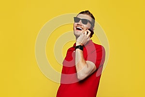 Portrait of a young cheerful man wearing red polo shirt and sunglasses having phone call on yellow backdrop.