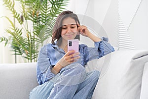 Portrait of a young cheerful woman sitting on a sofa in the living room at home, laughing with her mobile phone, smartphone in her