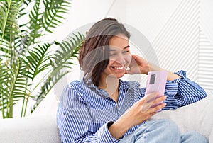 Portrait of a young cheerful woman sitting on a sofa in the living room at home, laughing with her mobile phone, smartphone in her