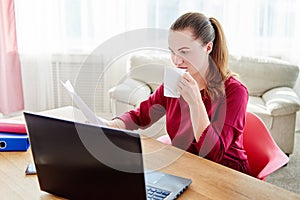 Portrait of young confident business woman sitting at wood desk, drinking coffee and reading documents in modern office.