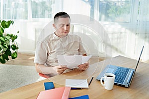 Portrait of young confident businessman sitting at wooden desk and working with documents and laptop in modern office, copy space