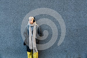 Portrait of young confident independent stylish hipster woman standing against black wall with wireless headphones listening music