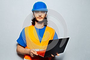 Portrait of young confident man, construction worker engineer wearing safety equipment, using laptop on grey background.