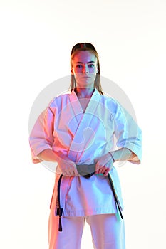 Portrait of young confident woman, female fighter in uniform with black belt isolated over white studio background.