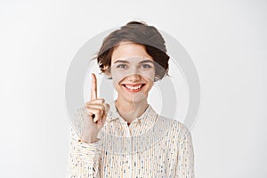 Portrait of young confident woman pointing finger up and smiling, showing promo logo, standing over white background in