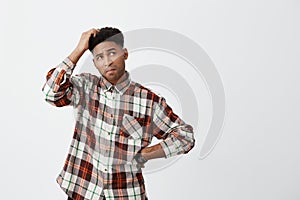 Portrait of young confused dark-skinned handsome man with afro hairstyle in checkered shirt holding head with hand