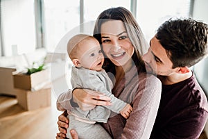 A portrait of young couple with a baby and cardboard boxes moving in a new home.