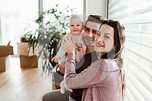 A portrait of young couple with a baby and cardboard boxes moving in a new home.