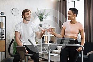 Portrait of young couple doing cardio on stationary bike and treadmill clinking bottles of water