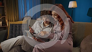 Portrait of young couple spending time together. Man and woman in pajamas sitting on the sofa, holding tablet and