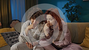 Portrait of young couple spending time together. Man and woman in pajamas sitting on the sofa, watching movie on tablet