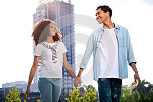 Portrait of young couple wearing casual clothing spending time together holding arms looking each other outdoors on
