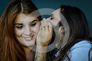 Portrait of a young couple where bearded man with long hair tells a funny secret to the woman with red hair and she smiles