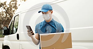 Portrait of young courier in mask standing near car holding carton box using smartphone.