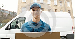 Portrait of young courier in mask standing near delivery car and holding carton box