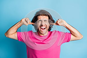 Portrait of young crazy wavy brunet hair man wear pink t-shirt fingers ears close eyes isolated on blue color background
