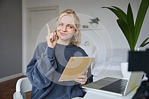 Portrait of young creative woman, content maker, sitting in room, working from home, using laptop, holding notebook