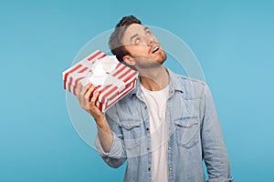 Portrait of young curious man in denim shirt holding gift box near ear and listening, guessing present