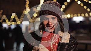 Portrait of young cute happy girl wearing winter hat holding present box, looking to camera and smiling. She standing in