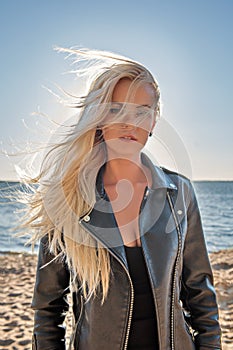 Portrait of a young cutie blond girl in a leather jacket with hair flying from the wind on the beach