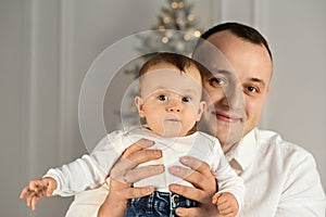 Portrait of a young dad with a baby in his arms, smiling near the Christmas tree