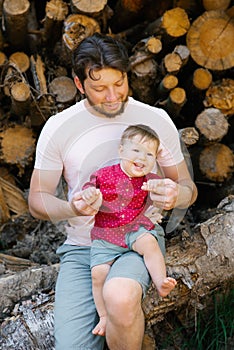 Portrait of a young dad and her child, whom she holds in her arms, sitting on logs in the forest in summer