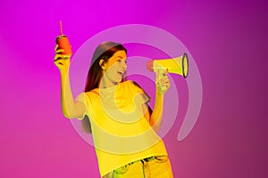 Portrait of young delightful girl posing, shouting in megaphone isolated over pink background in neon light