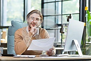 Portrait of a young designer, architect, engineer sitting in an office, working at a computer. He holds his chin with
