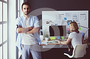 Portrait of young designer in front of laptop and computer while working. Assistant using her mobile at background.