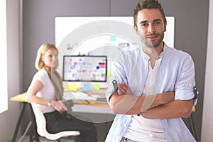 Portrait of young designer in front of laptop and computer while working. Assistant using her mobile at background.