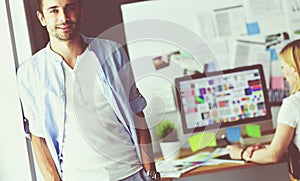 Portrait of young designer in front of laptop and computer while working. Assistant using her mobile at background.