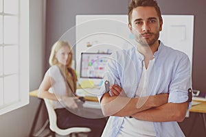 Portrait of young designer in front of laptop and computer while working. Assistant using her mobile at background.