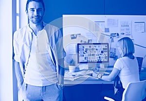 Portrait of young designer in front of laptop and computer while working. Assistant using her mobile at background.