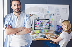 Portrait of young designer in front of laptop and computer while working. Assistant using her mobile at background.