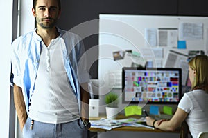 Portrait of young designer in front of laptop and computer while working. Assistant using her mobile at background.