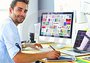 Portrait of young designer sitting at graphic studio in front of laptop and computer while working online.