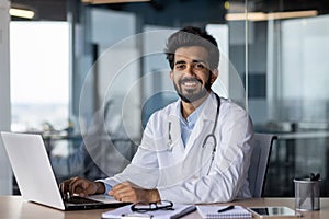 Portrait of a young doctor of Indian origin working in the office using a laptop, sitting at the table and smiling at